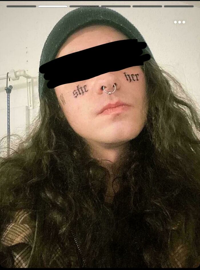 Person with long curly hair and a nose ring showing a face tattoo with words in gothic script, a truly awful tattoo example.