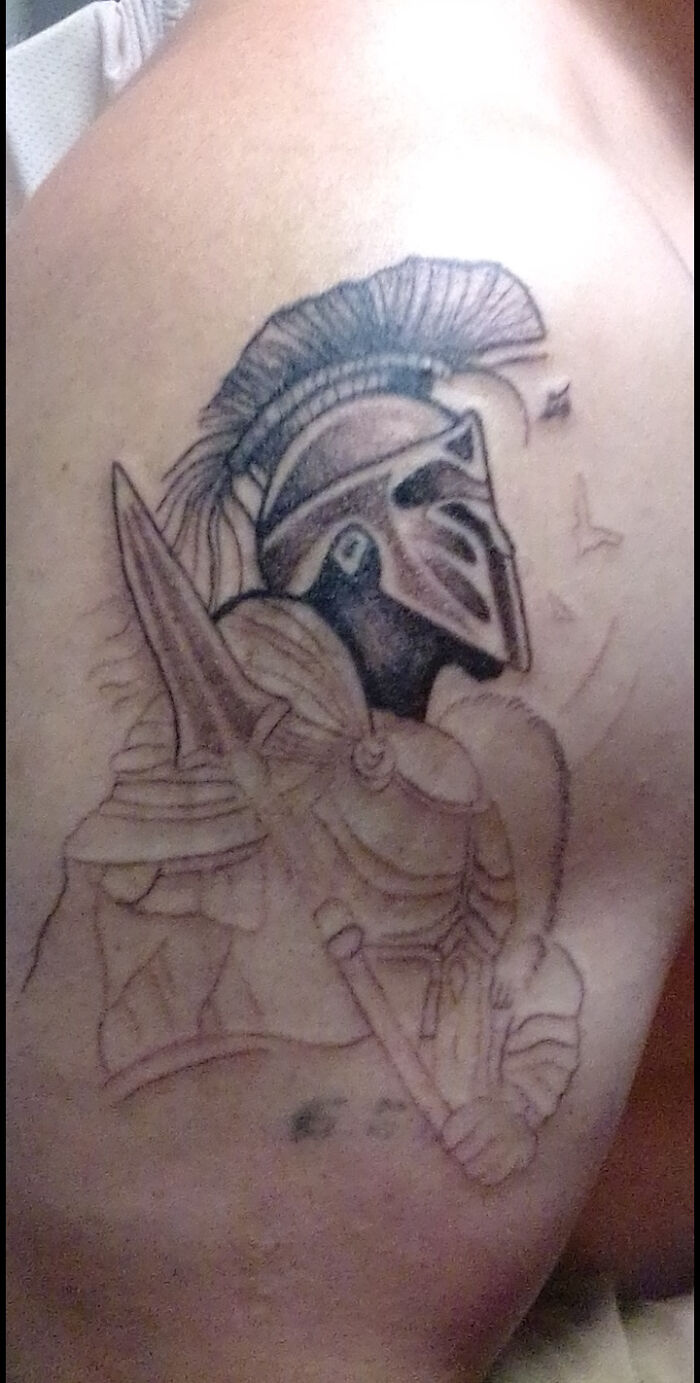 Tattoo of a poorly detailed medieval knight in armor with a large helmet and sword on a person's upper shoulder.