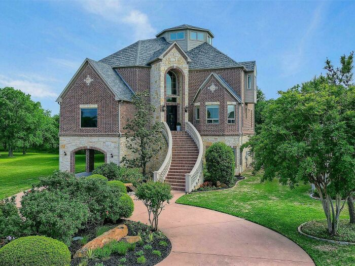 A large brick mansion with a grand curved staircase leading to the entrance, surrounded by green lawns and trees.