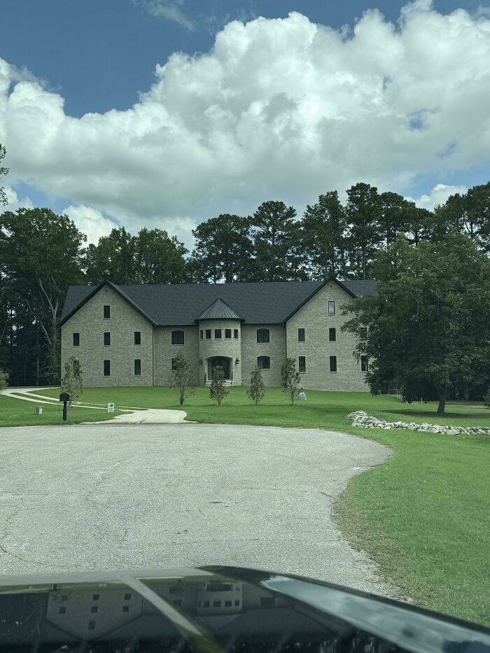 A large modern mansion with a symmetrical design, dark roof, and a prominent circular entry, surrounded by trees.