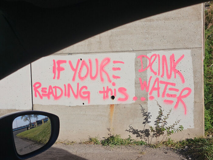 Graffiti with poor sign design on a concrete wall reads 'If You're Reading This... Drink Water', a confusing message.