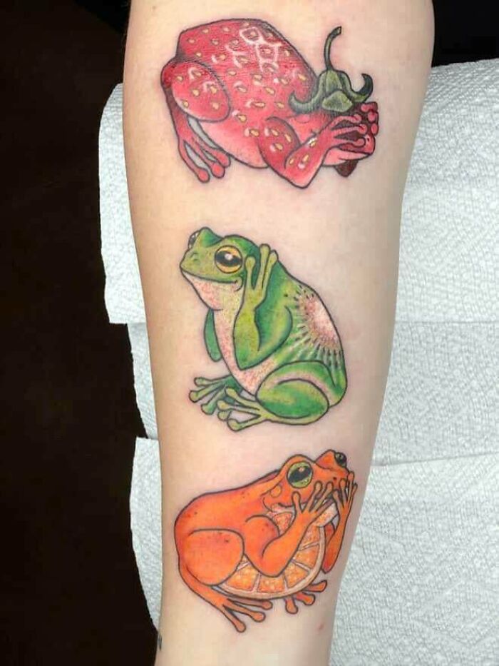 Three colorful, stunning tattoos of frogs disguised as fruits: a strawberry, a regular green frog, and an orange, illustrating stunning tattoos.