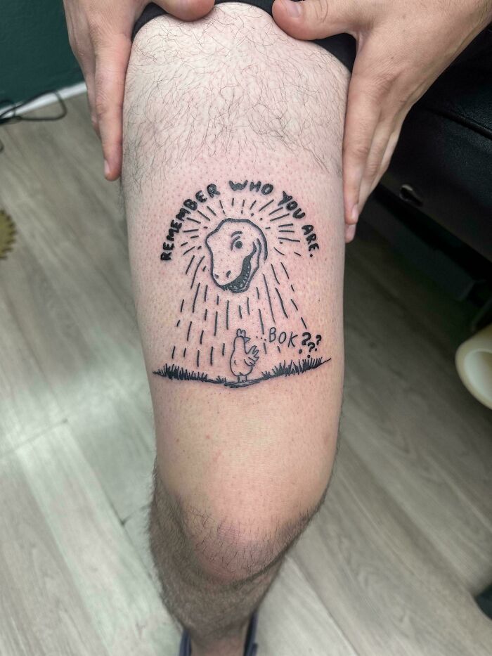 A person displays a unique, humorous tattoo on their leg featuring a chicken looking up at a shining rock with text.