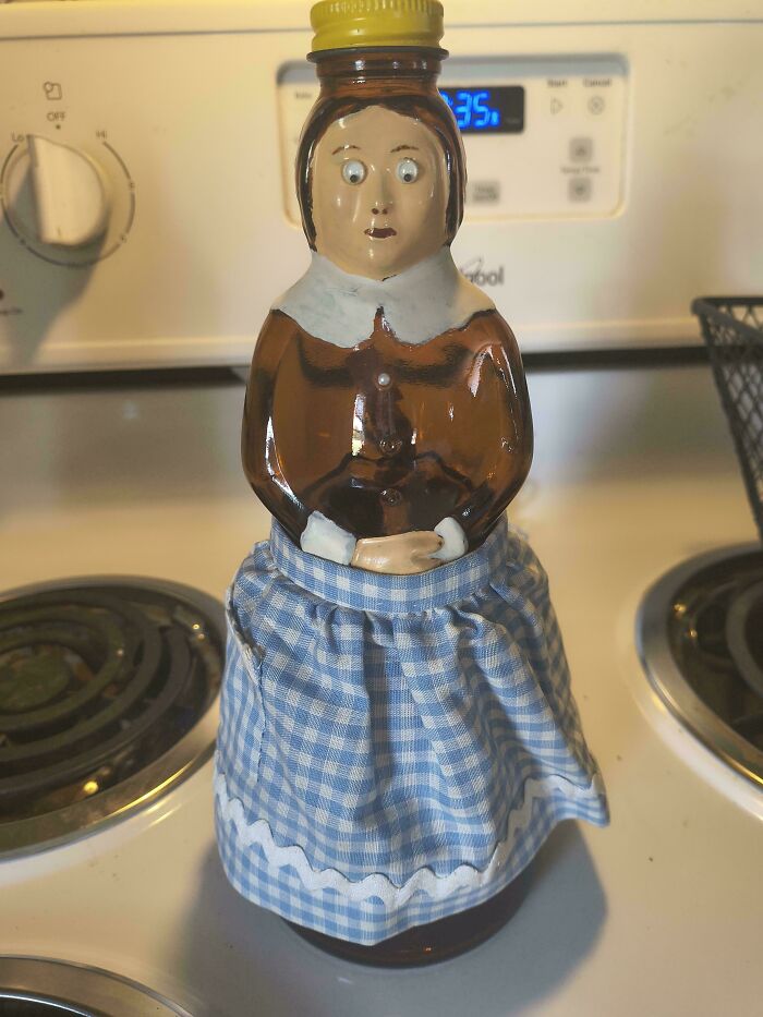 A DiWHY DIY glass syrup bottle shaped like a woman in a blue gingham skirt, with wide, painted eyes, on a stove.