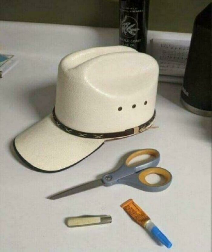 A cowboy hat converted to a baseball cap with scissors, knife, and glue on a table, a DiWHY craft gone wrong.