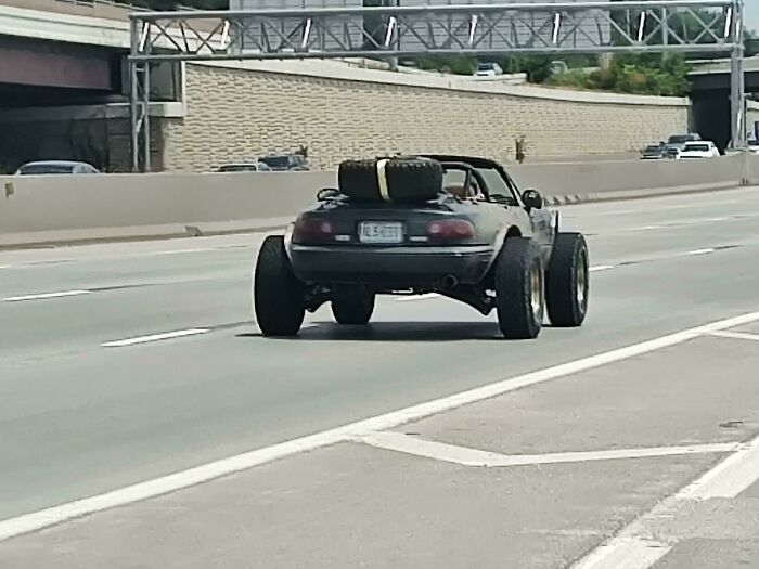 A black "DiWHY" car with oversized tires and a spare on the trunk, driving on a highway.