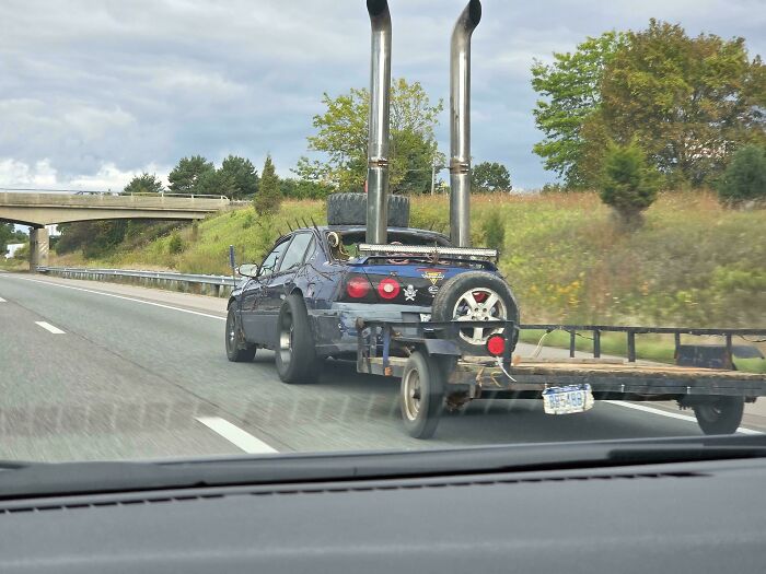 A heavily modified DiWHY car with tall exhaust pipes, spikes, and a trailer, driving down a highway.