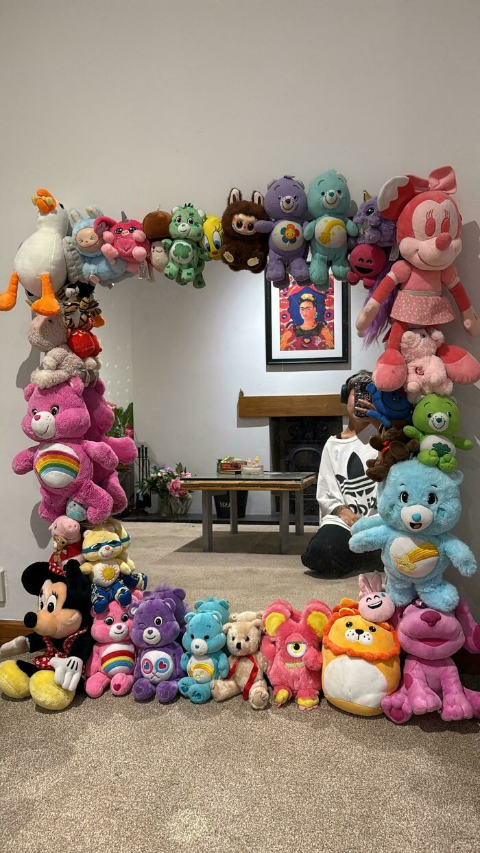 A DiWHY project: a mirror frame covered in various colorful stuffed animals, reflecting a person in a white room.