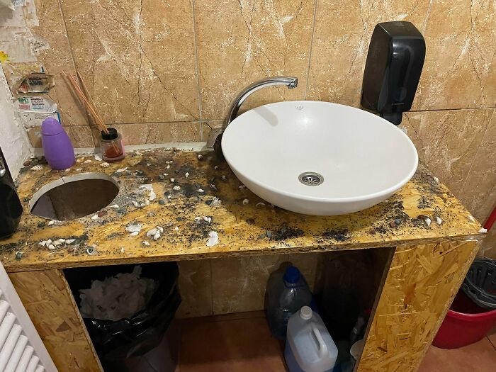 A messy "DiWHY" bathroom with a white sink and a particle board counter covered in debris.