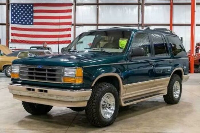 A classic dark green and tan '90s Ford Explorer SUV with an American flag in the background, a nostalgic '90s pic.