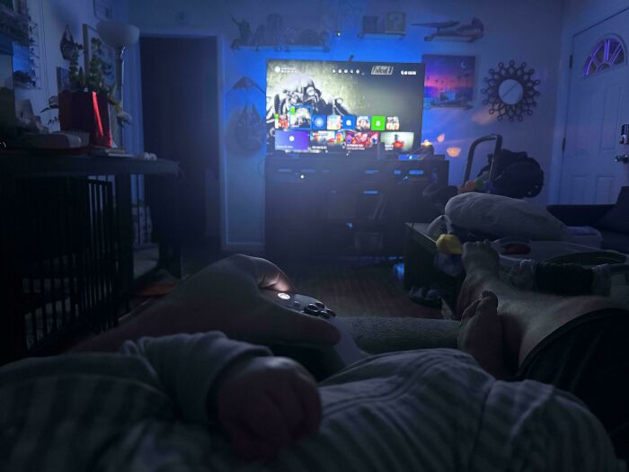 A dad playing video games with a baby sleeping on his chest, showcasing feel-good posts for dads.
