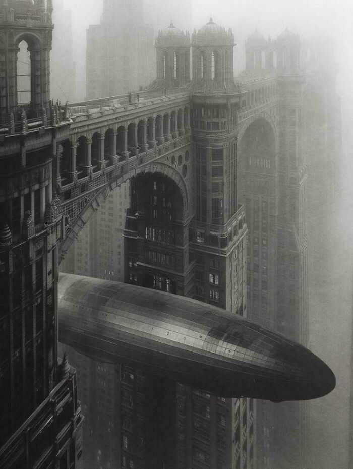 A monochrome, foggy cityscape with ornate buildings connected by bridges, an impressive architecture, and a large airship.
