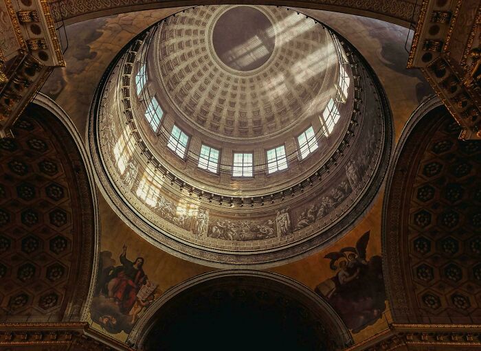 A grand architectural dome with natural light streaming through windows, highlighting intricate details and paintings, an impressive building.