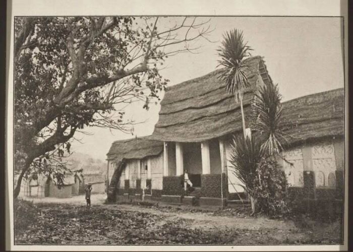 Sepia-toned photo of impressive architecture: thatched-roof buildings, tall palms, and a tree with bare branches.