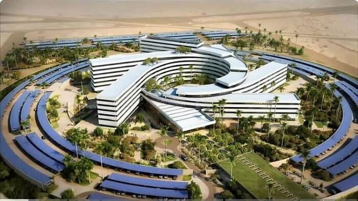 An aerial view of a modern, curved building complex surrounded by solar panel arrays and palm trees in a desert landscape. Architecture lovers.