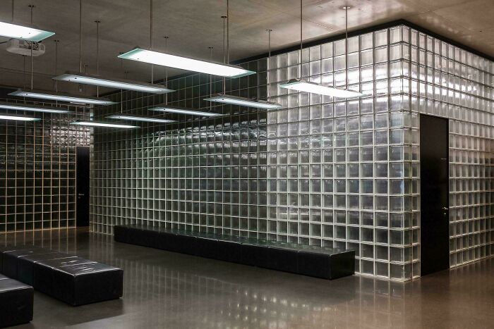 Modern architecture with a glass block wall, black benches, and linear ceiling lights in a minimalist building.