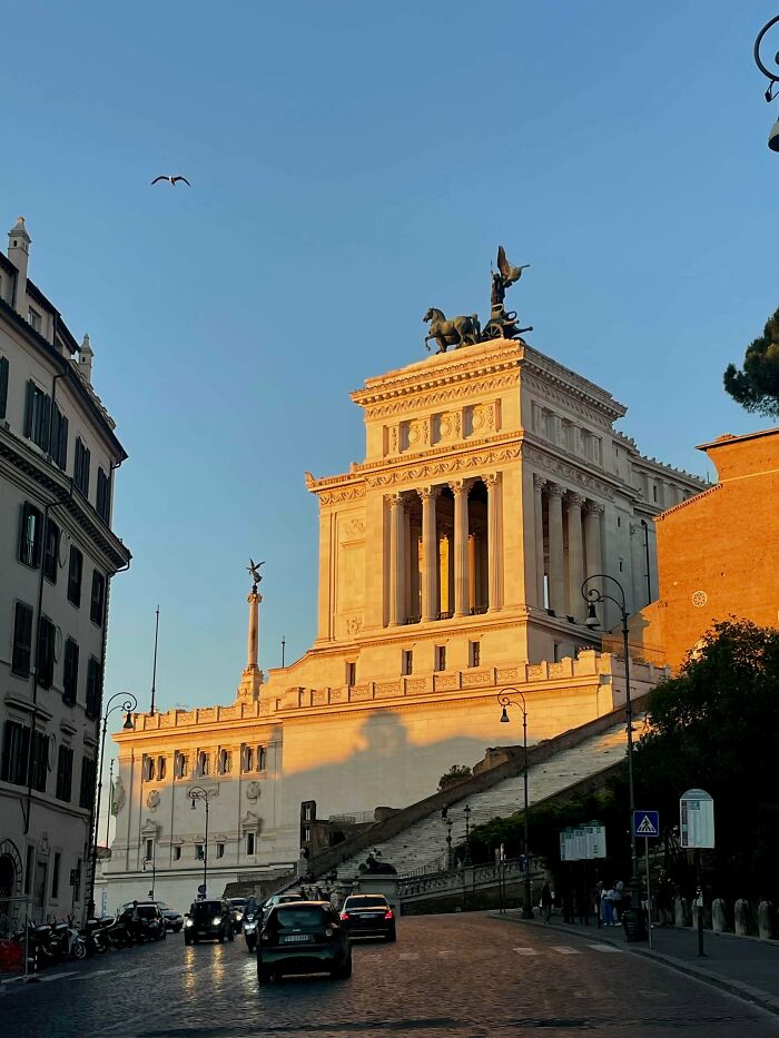 A grand building, bathed in golden hour light, showcasing impressive architecture. Cars drive on a street below.