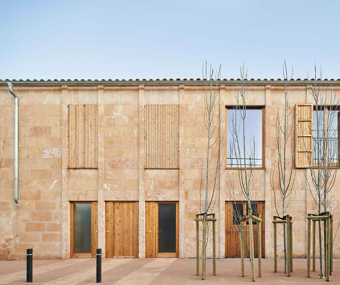 An impressive architecture building with a stone facade, wooden panels, windows, and doors, complemented by bare young trees.