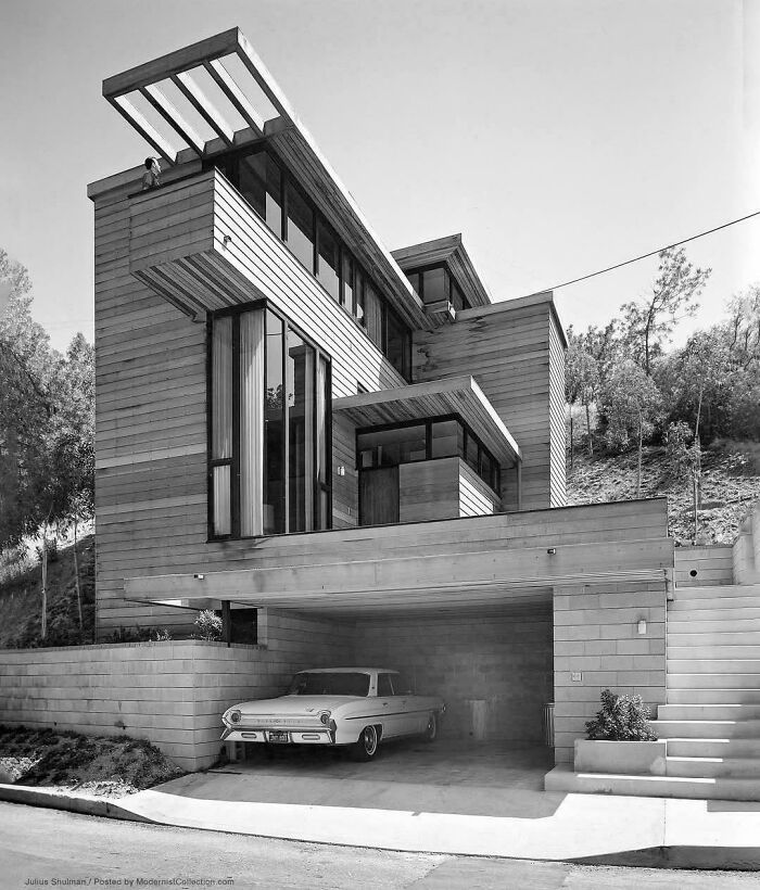 Black and white photo of an impressive modernist building with wood siding, large windows, and an integrated garage. Architecture lovers will appreciate the design.