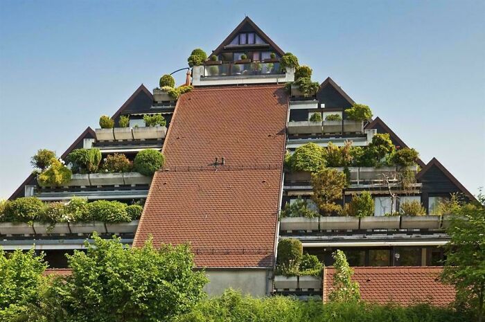 A striking pyramid-shaped building with terraced balconies overflowing with lush green plants under a clear blue sky. Architecture lovers will be impressed.