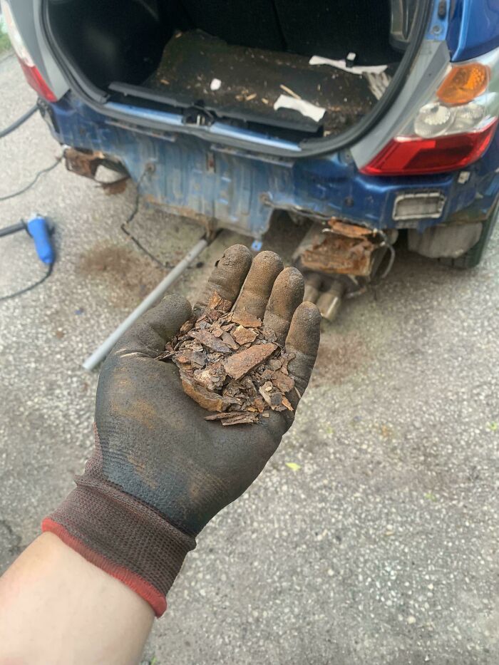 Mechanic’s glove holding rust flakes in front of a heavily rusted car bumper being repaired outdoors.
