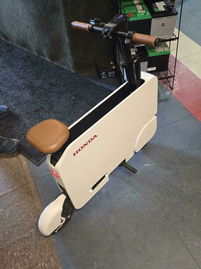 Compact white Honda electric scooter with brown seat and handlebars, shown indoors on tiled floor by mechanics.