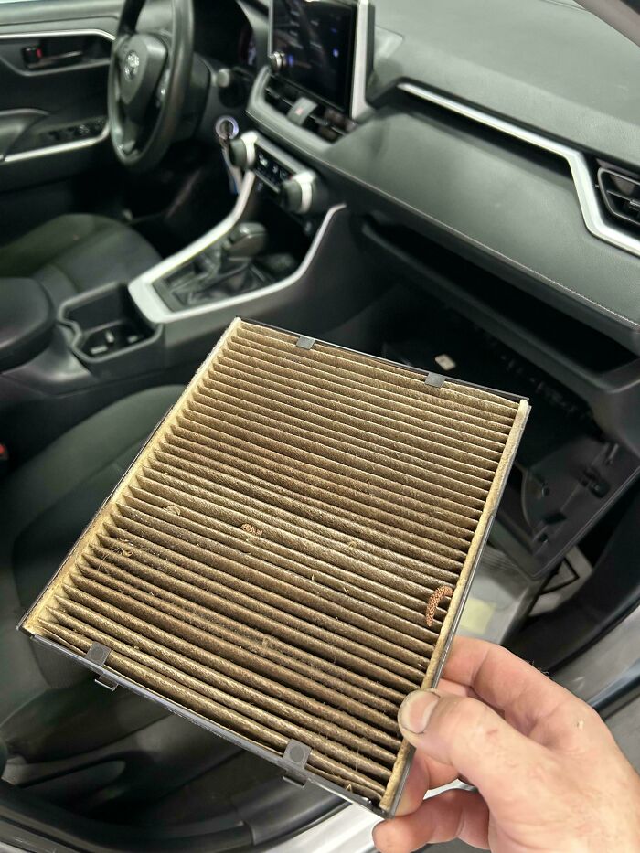 Hand holding a dirty car air filter inside a vehicle, showing a mechanic's work and car maintenance issue.