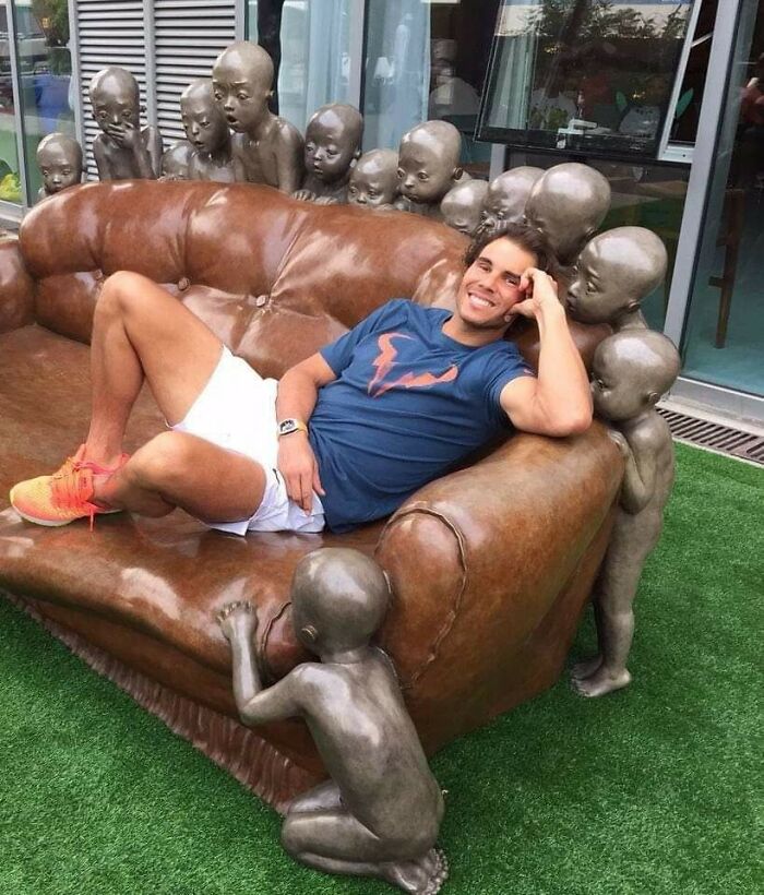 Man relaxing on a brown leather couch surrounded by creepy child sculptures in a bizarre and ridiculous piece of furniture.