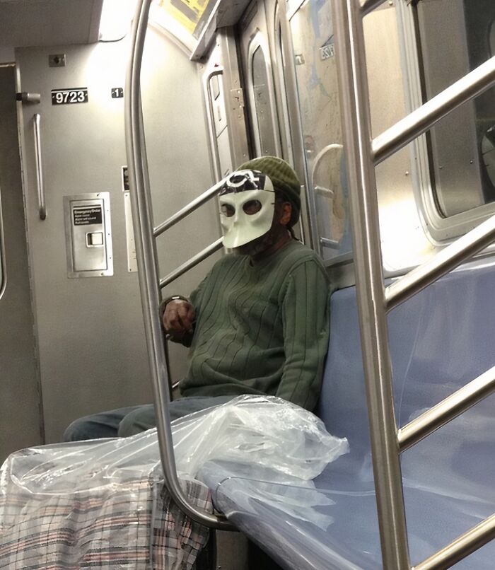 Person wearing a white mask and green sweater sitting alone on a NYC subway bench with bags nearby.