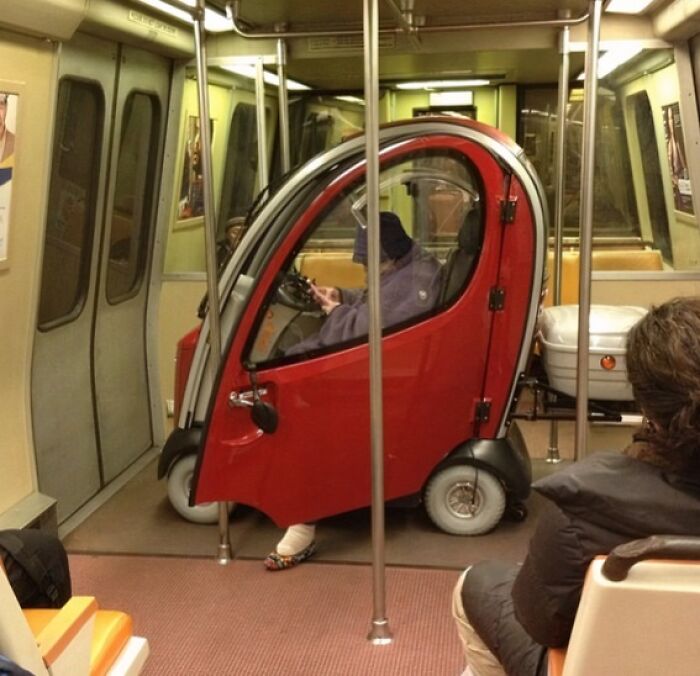Person riding a small enclosed mobility scooter inside a NYC subway car, surrounded by seated passengers.
