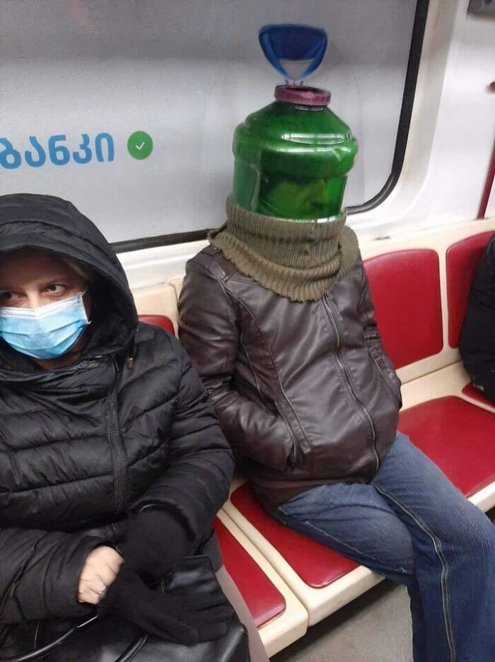 Person wearing a large green water jug on their head sitting next to a masked passenger on a subway train.