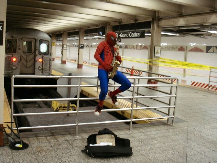 Person dressed as Spider-Man playing saxophone on a railing at a NYC subway station near a train.