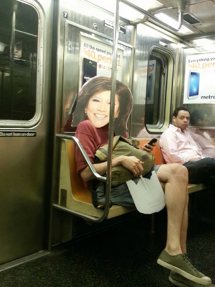 Person on NYC subway wearing large cutout mask of a smiling face while holding a bag and sitting next to a passenger.