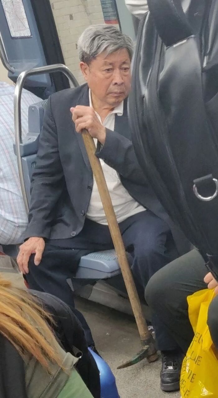 Elderly man holding a pickaxe while sitting on a seat inside a crowded NYC subway car.