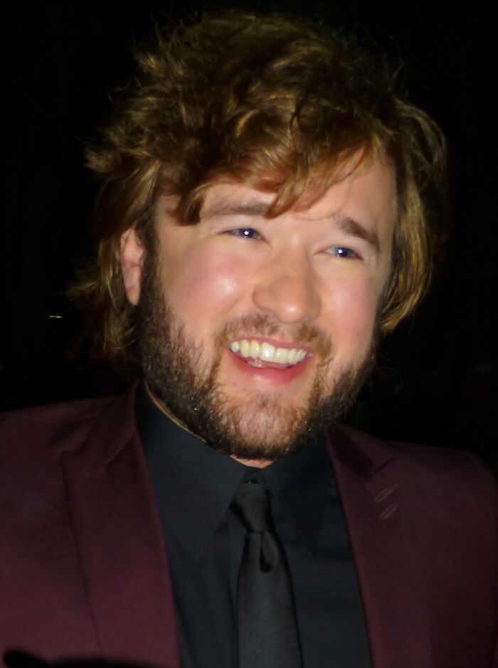 Bearded man smiling in a dark setting wearing a maroon jacket and black shirt, representing iconic actors who dominated Hollywood.