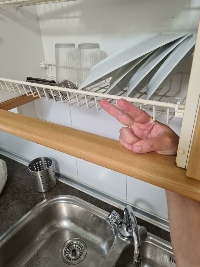 Hand showing a peace sign inside a kitchen cabinet with dishes drying above a stainless steel sink.