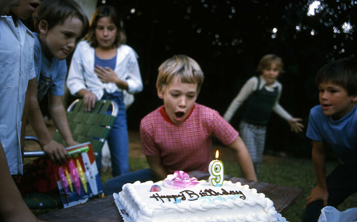 A boy blowing out his 9th birthday candle with excited friends around, showcasing an Accidental Renaissance look.