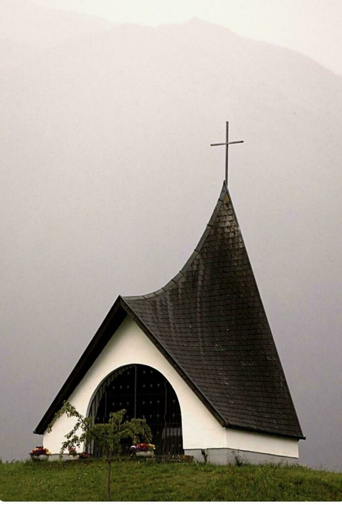 Small, beautiful architecture of a chapel with a dark, pointed roof and cross, set against a misty mountain background.