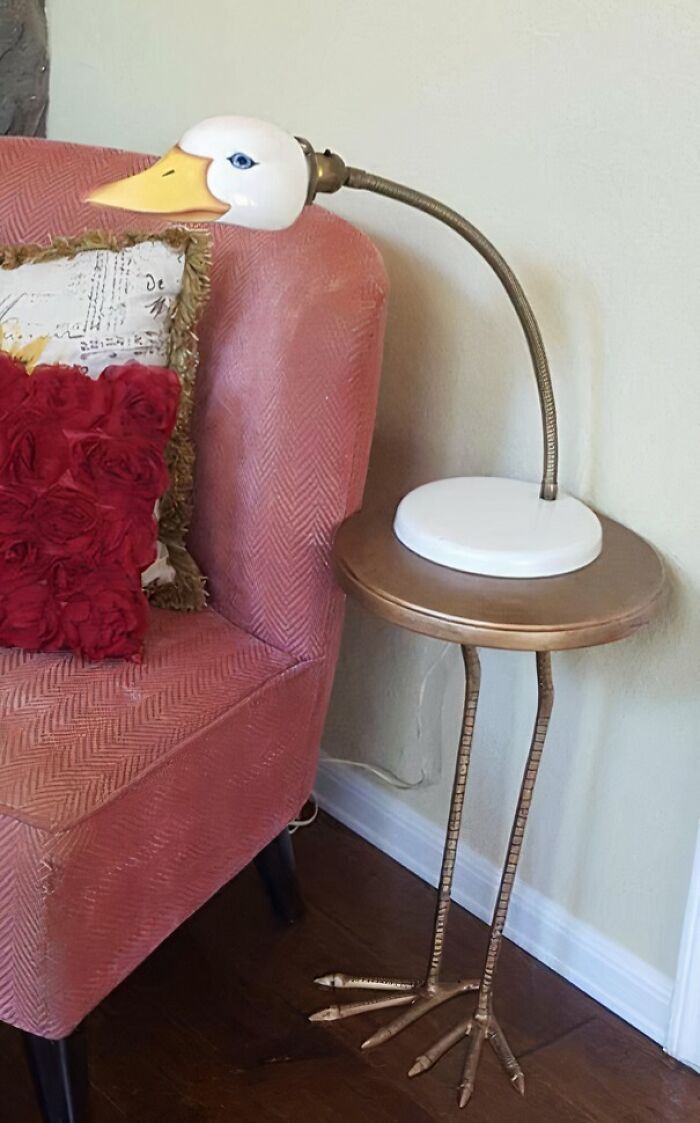 Ridiculous piece of furniture shaped like a duck with long legs, a lamp head, and a small round table beside a pink chair.