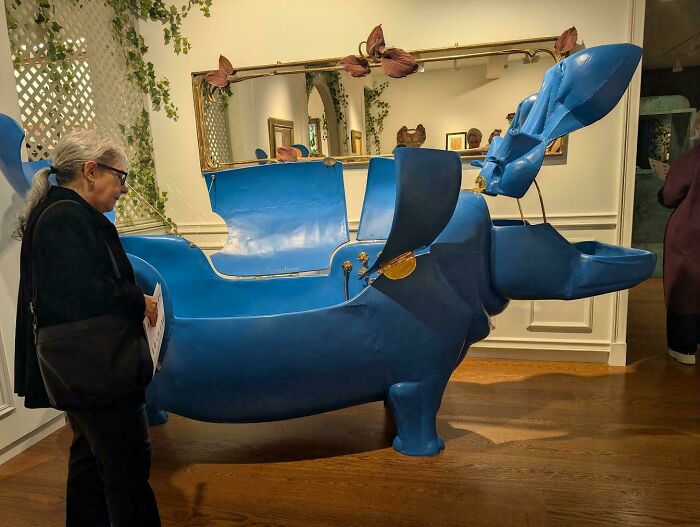 A woman examining a large, blue, dog-shaped piece of furniture in a well-lit room with wooden floors.