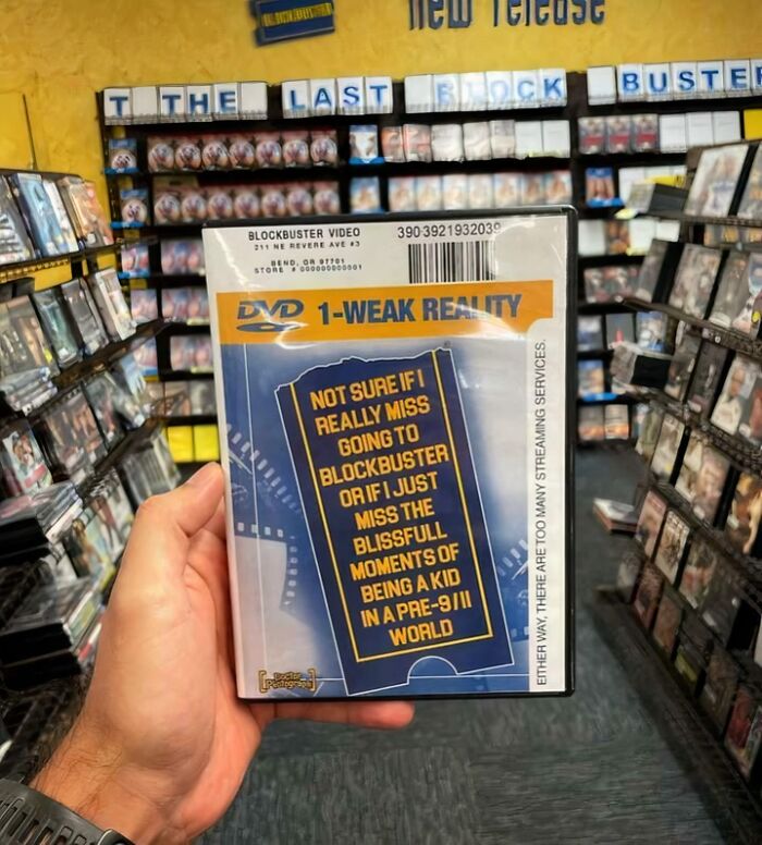 Hand holding DVD case with nostalgic Blockbuster reference in video rental store filled with ’90s movie shelves and memos.