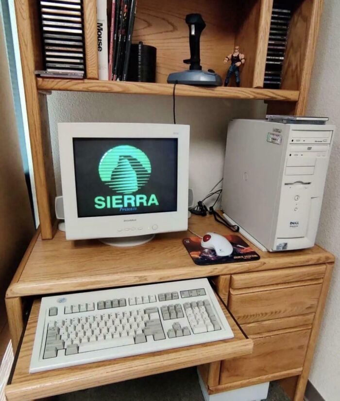 Vintage 90s computer setup with CRT monitor showing Sierra logo, classic keyboard, and joystick on wooden desk in home office.
