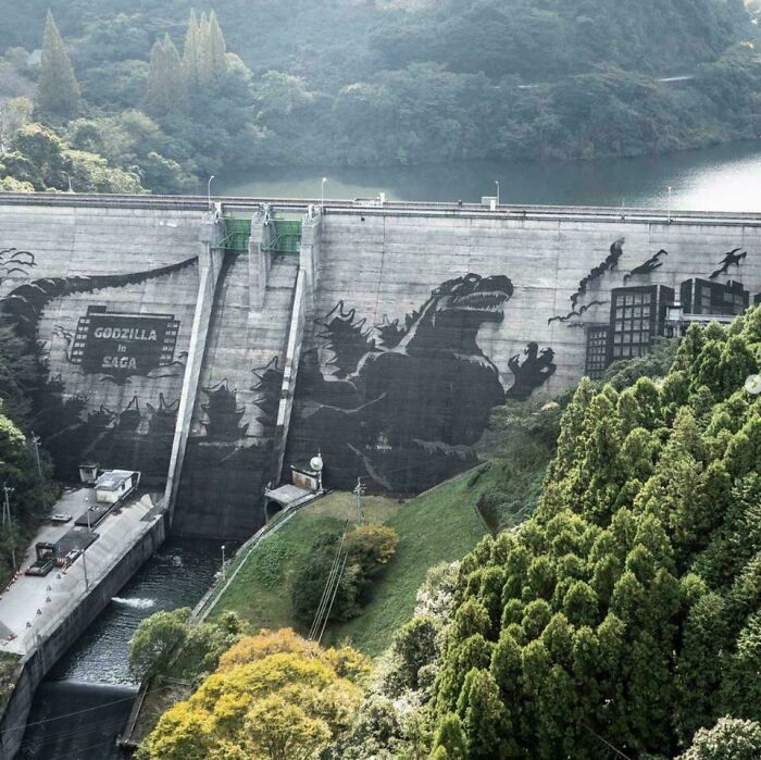 Mural of Godzilla painted on a large dam wall surrounded by lush green trees in an incredible reality scene.