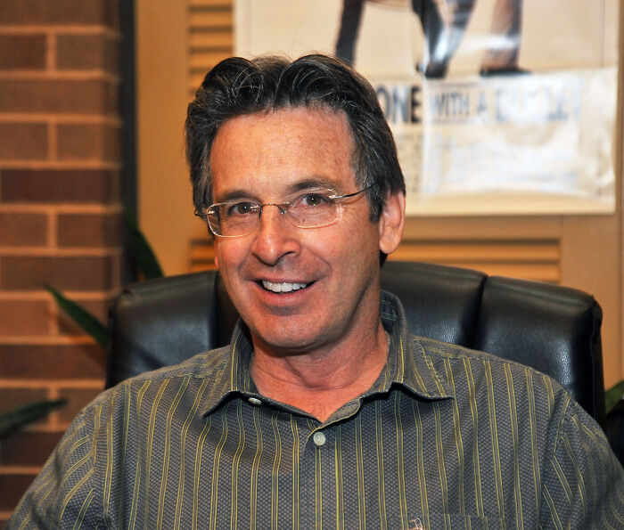 Robert Carradine smiling, seated indoors wearing glasses and a striped shirt, linked to Disney's Lizzie McGuire controversy.