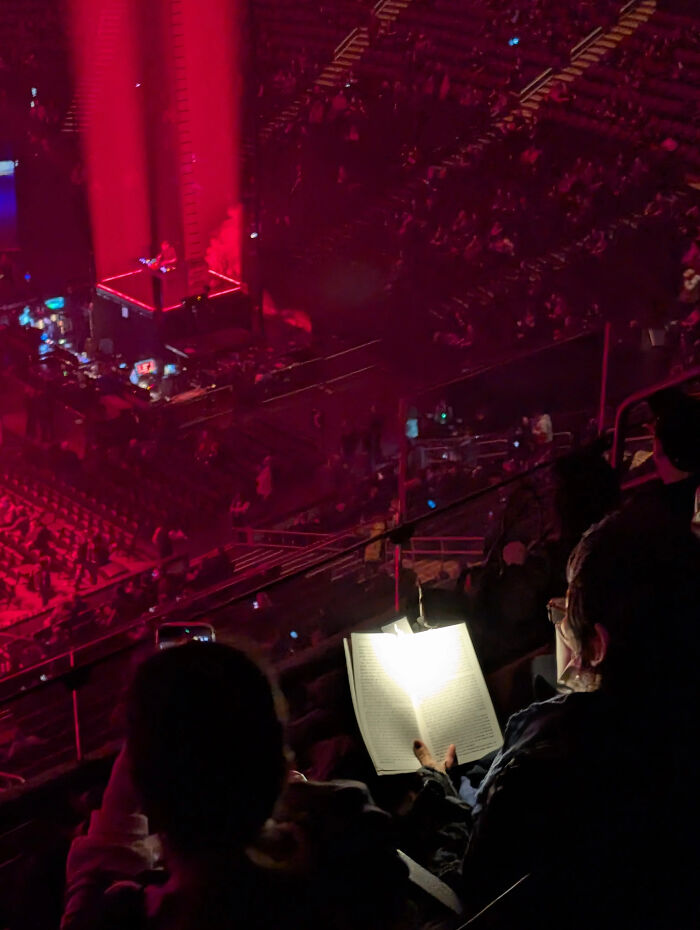 An audience member reading a book under a bright light during a concert, in an Accidental Renaissance style.