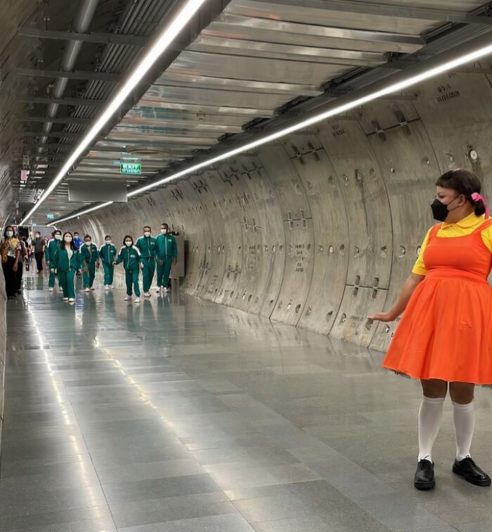 Person in bright orange dress and mask posing in NYC subway tunnel with a group of people in green tracksuits walking behind.