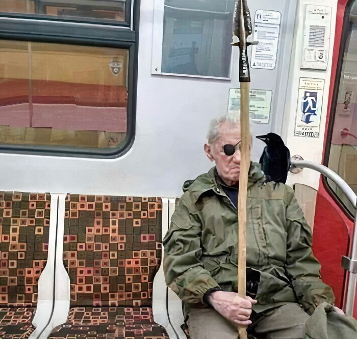 Elderly man with an eyepatch holding a spear and a black bird perched on his shoulder on a NYC subway train.