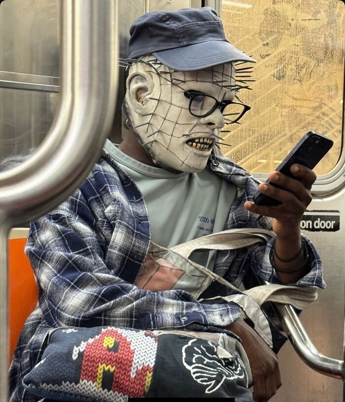 Person wearing a spiked mask and glasses, sitting on a NYC subway and looking at a smartphone.
