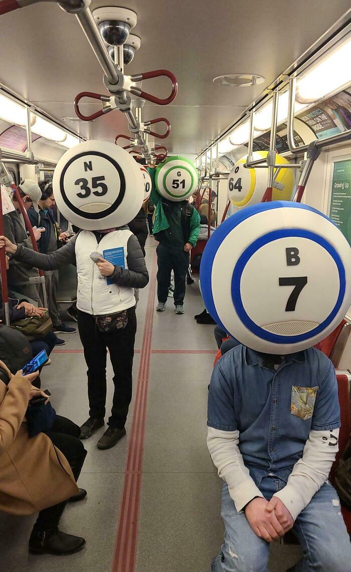 People wearing oversized numbered subway token headpieces in a crowded NYC subway car, creating a weird subway moment.