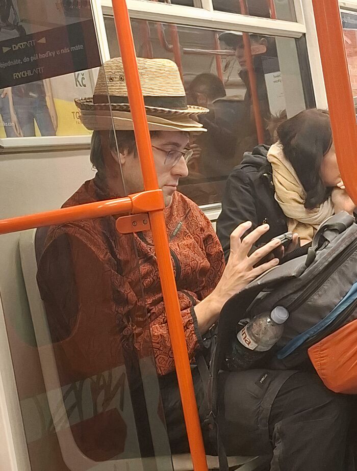 Man wearing multiple straw hats and an orange patterned jacket, using phone on NYC subway amid weird subway moments.